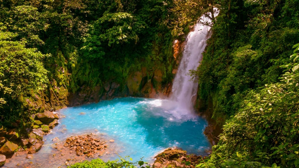 A waterfall cascades into a vibrant blue pool, surrounded by lush green vegetation and rocks in a forest setting—the perfect scene to experience when exploring during the best time of year to visit Costa Rica.